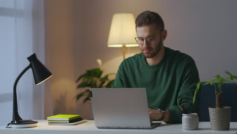 man with glasses is sitting in front of laptop thinking and typing on keyboard working at evening at home freelancer writer or journalist