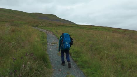 A-hiker-with-a-blue-backpack-walks-up-a-grassy-hillside-on-a-cloudy-day