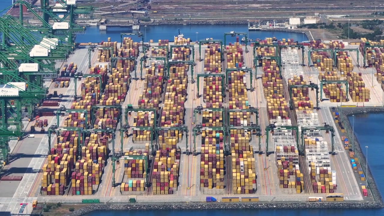 Aerial view of a busy port with numerous containers. Ships are docked, and cargo handling activities are ongoing. Delays in cargo delivery are evident due to high congestion levels.