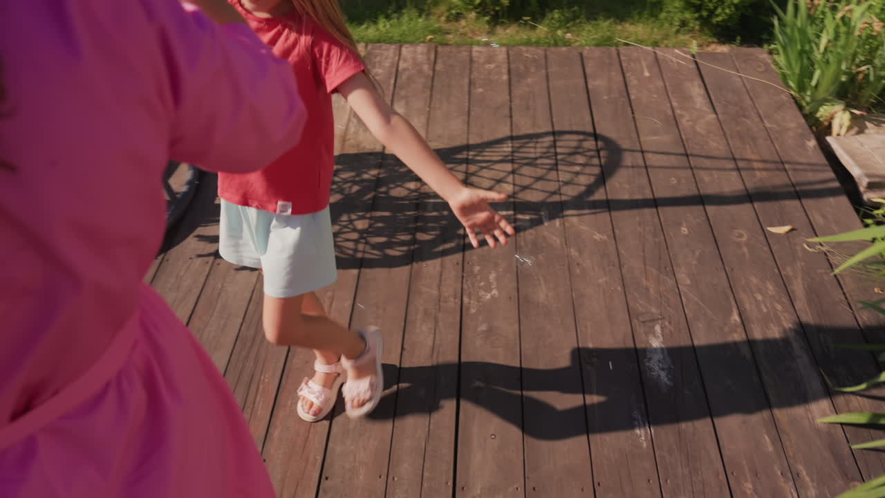 Child Chases Shimmering Soap Bubbles, Children Enjoy Outdoor Fun Blowing Bubbles On Sunlit Porch, Playful Summer Scene With Mother Blowing Sparkling Bubbles For Daughter To Chase Outdoors