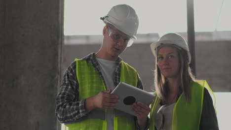 Dos-Ingenieros,-Un-Hombre-Y-Una-Mujer-Con-Cascos-Blancos-Y-Una-Tableta-En-El-Sitio-De-Construcción,-Condenan-El-Plan-De-Construcción-Del-Edificio