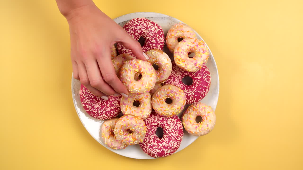 Plate full of colorful Donuts in pink and white on light yellow background. Female Hand putting small doughnut on big ones. Top view, filmed in Prores.