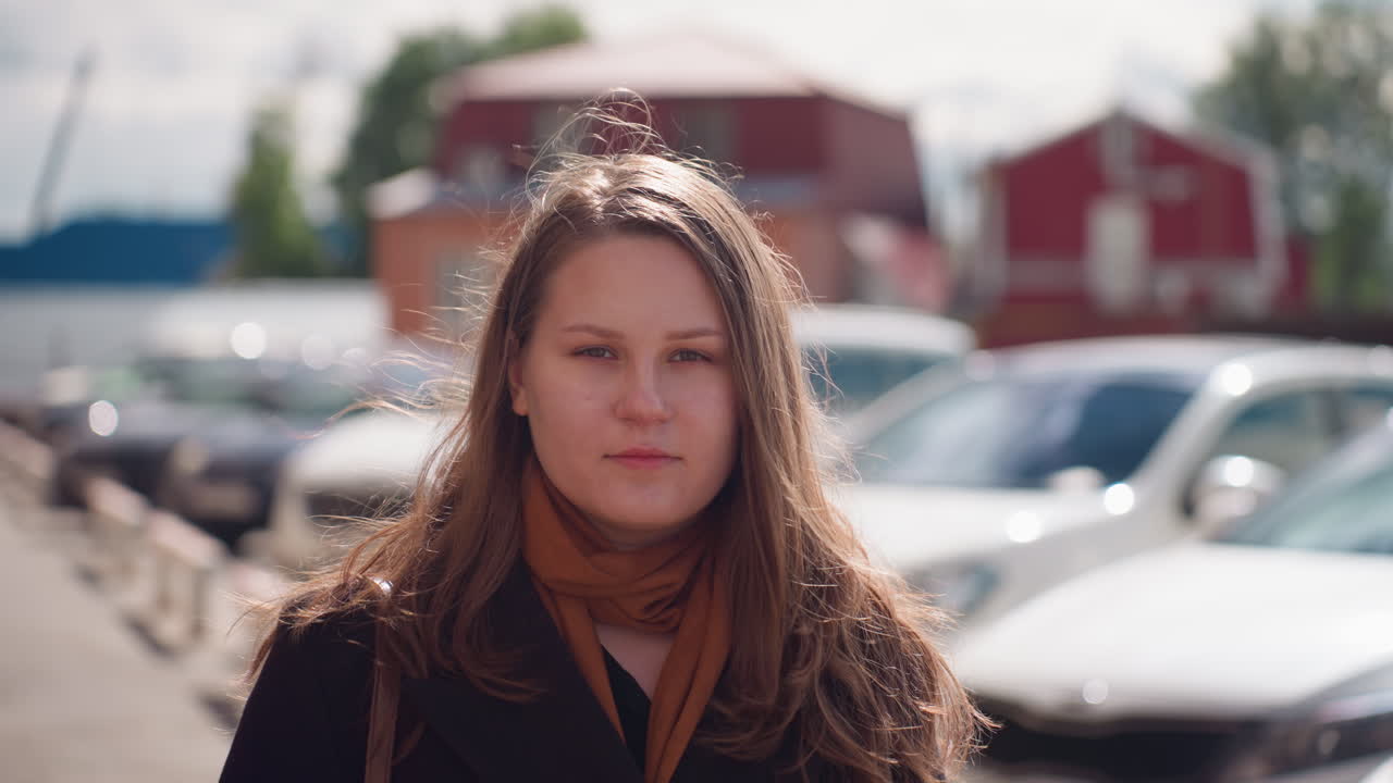 Student in coat adjusts sunglasses under bright sun as breeze lifts hair, Blurred city parking lot and brick building behind create calm, confident mood in soft afternoon light of everyday life