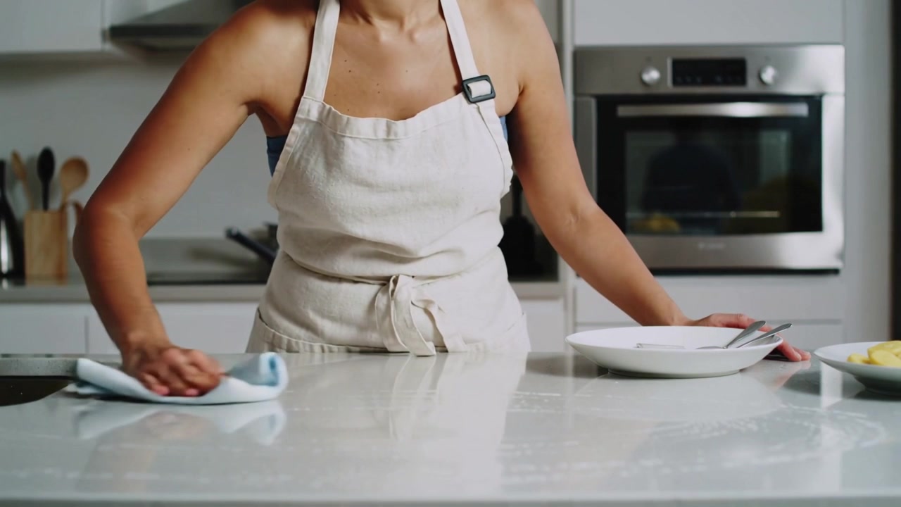 Mulher de avental limpando a cozinha depois de cozinhar, segurando um pano azul e um prato com talheres.