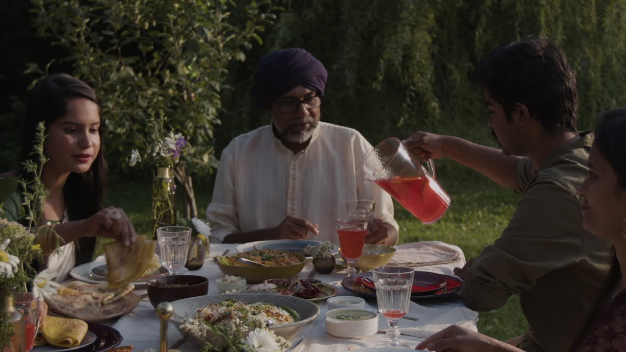 A family enjoys an outdoor meal with traditional Indian dishes and drinks in a garden setting