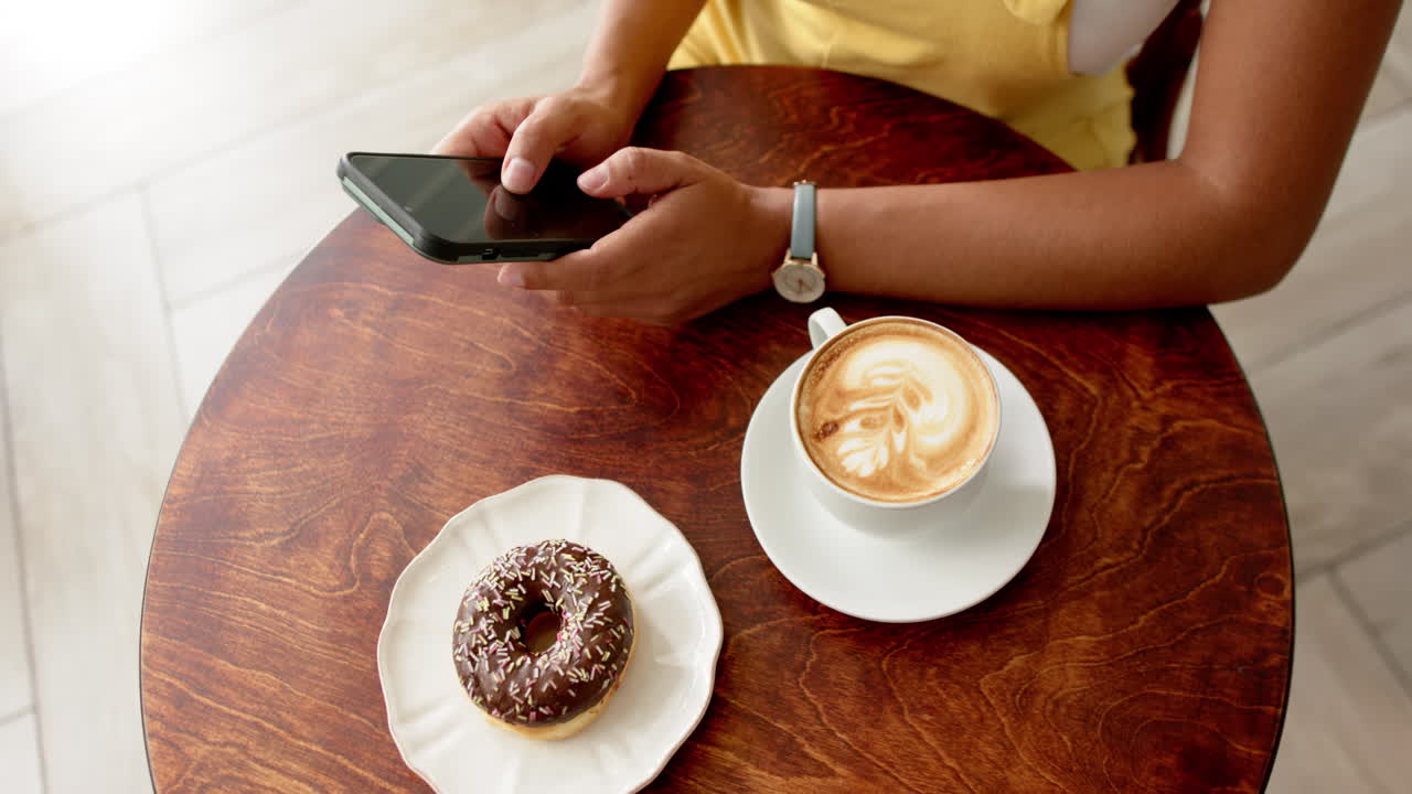 Young biracial woman using a smartphone at a cafe table with a doughnut and coffee in a cafe