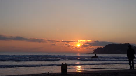 silueta de un fotógrafo tomando una foto de un surfista sosteniendo una tabla de surf mientras caminaba hacia el mar en japón contra las olas con un glorioso paisaje de puesta de sol en el fondo - plano general
