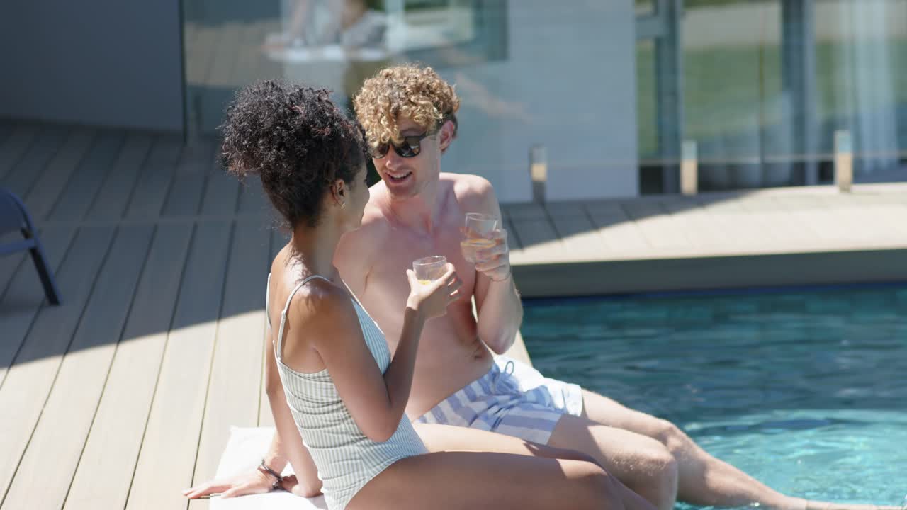 Diverse couple exchanging eye contact, clinking glasses, sharing on rooftop pool deck in swimwear