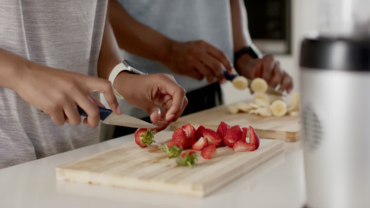 Coppia che affetta fragole e banane sul piano di lavoro della cucina con i coltelli, preparando la frutta per il frullatore.
