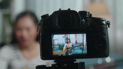 close up of a camera monitor recording asian woman reading comments on laptop and speaking while live stream playing a guitar at home