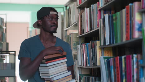 Student in casual clothing holding stack of books walks through library aisle, scanning shelves with focused expression, searching for more reading material among rows of colorful books