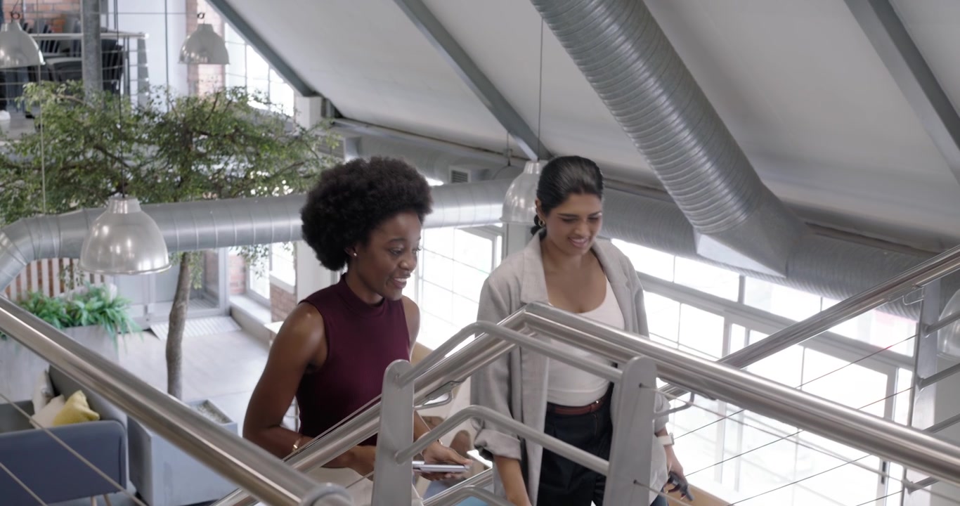 African American, Indian female coworkers walking in loft, checking smartphone, ascending staircase