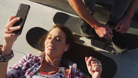 happy caucasian woman using smartphone and man tying shoe at a skatepark