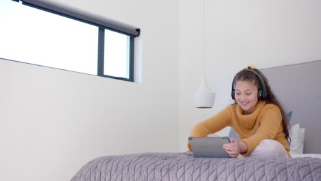 happy biracial girl sitting on bed, using tablet and headphones in sunny bedroom, copy space