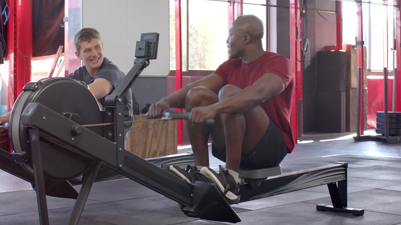 Diverse male friends at gym adjusting straps then rowing together on rowing machines for fitness