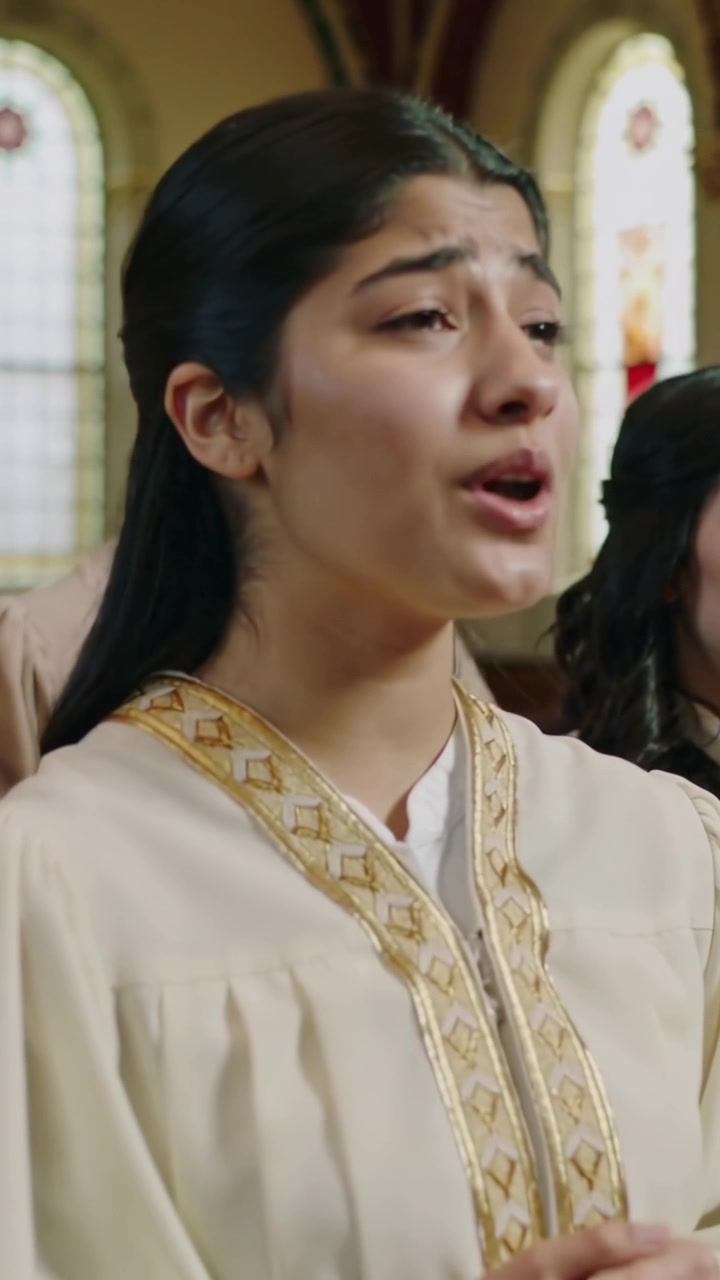 Vertical video: Singing choir member in cream robe gold trim for worship by stained-glass pullback