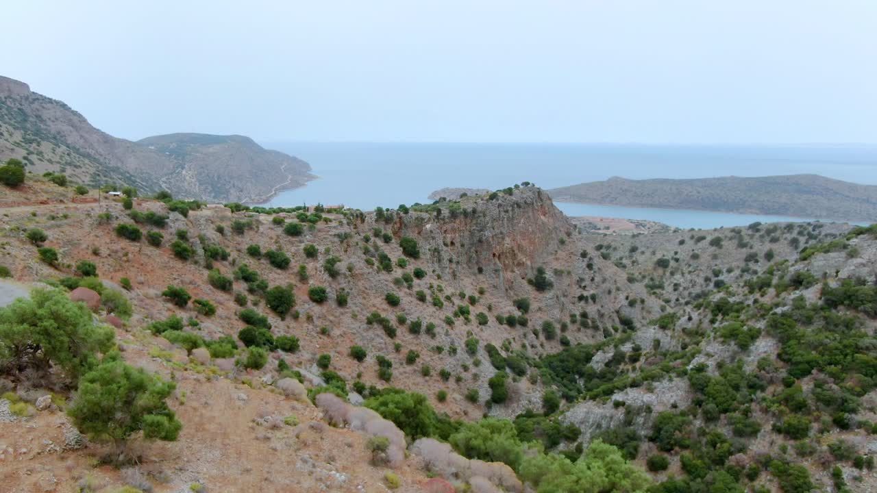 Barren dry coastal landscape on stony hills with isolated bushes with a road and view of the sea in the distance. Drone shot