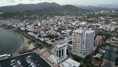 Puerto-Deportivo-De-La-Ciudad-De-Santa-Marta-Aérea,-Colombia,-Rompeolas-Y-Paisaje-Urbano-Horizonte-Montaña-Sierra-Nevada-Y-Hotel-De-Lujo-En-El-Rodadero,-Vista-Aérea-De-Veleros