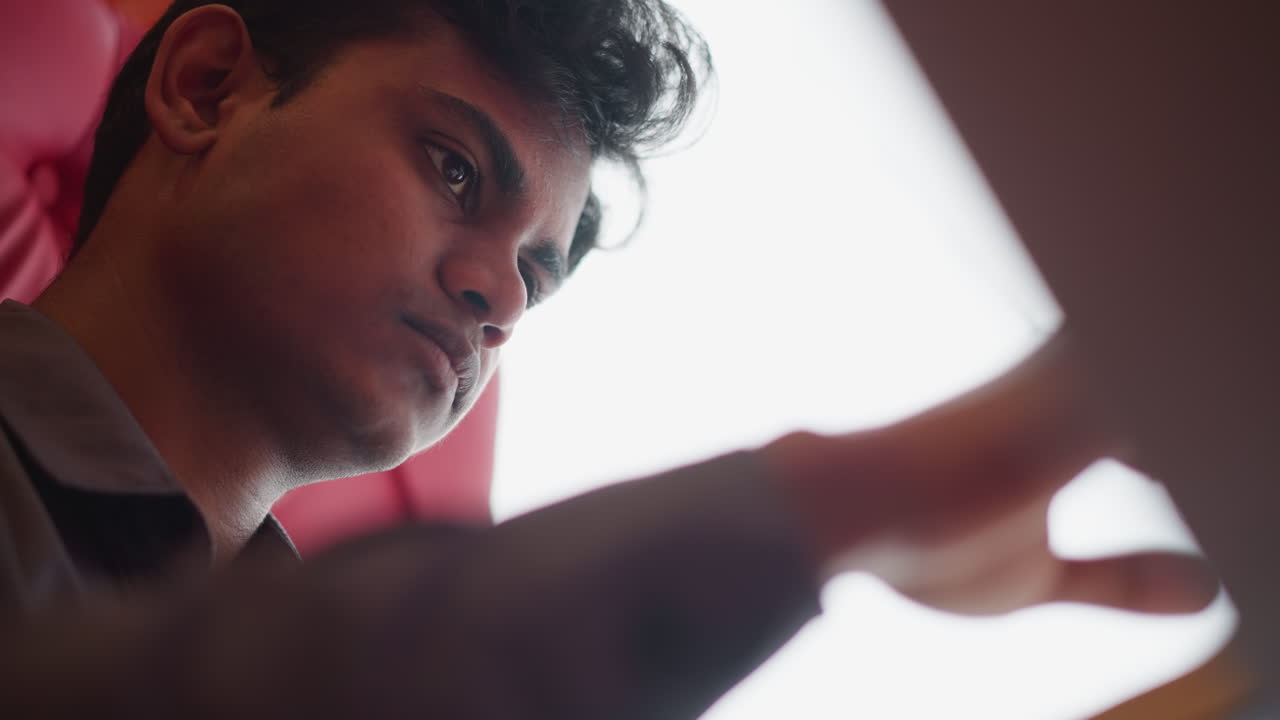 Close-up side view of male sitting against red cushioned seat, deeply focused while reading, with soft lighting from window highlighting his thoughtful expression, capturing quiet indoor study
