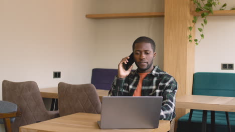 Man-Talking-On-Cellphone-While-Working-With-Laptop-Computer-Sitting-At-Table-At-Cafe