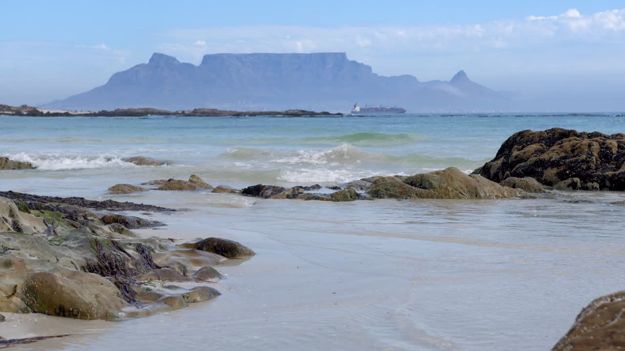 Waves gently splash over rocks in foreground with table mountain and a ship in the distance