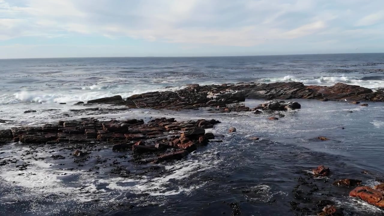Ampio paesaggio marino costiero con rocce scure e frastagliate, onde che si infrangono e un orizzonte calmo sotto una luce diurna soffusa e nuvolosa.