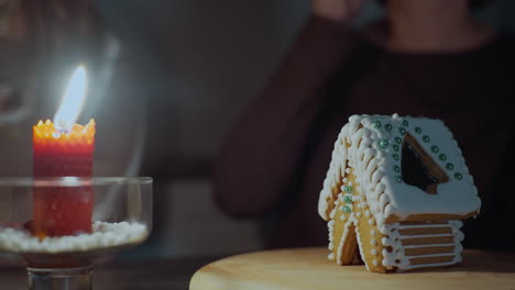 close-up of red candle burning beside intricately decorated gingerbread house on wooden tray, warm glow illuminates cozy scene as slightly blurred people in background raise cups in a toast