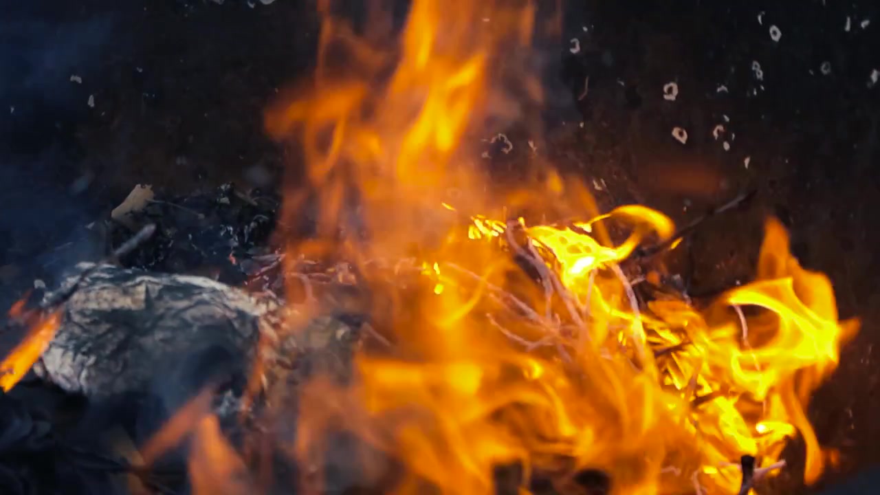 Fiamme arancioni brillanti e fumo che consumano spazzatura e rami secchi all'interno di un braciere di metallo scuro. Un piccolo falò che fornisce calore e luce in un giardino durante una fresca serata all'aperto