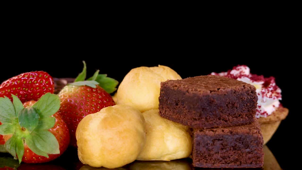 A colorful assortment of brownies, cream puffs, tarts, and fresh strawberries rotates smoothly under bright studio lighting against a seamless black background, highlighting textures and details