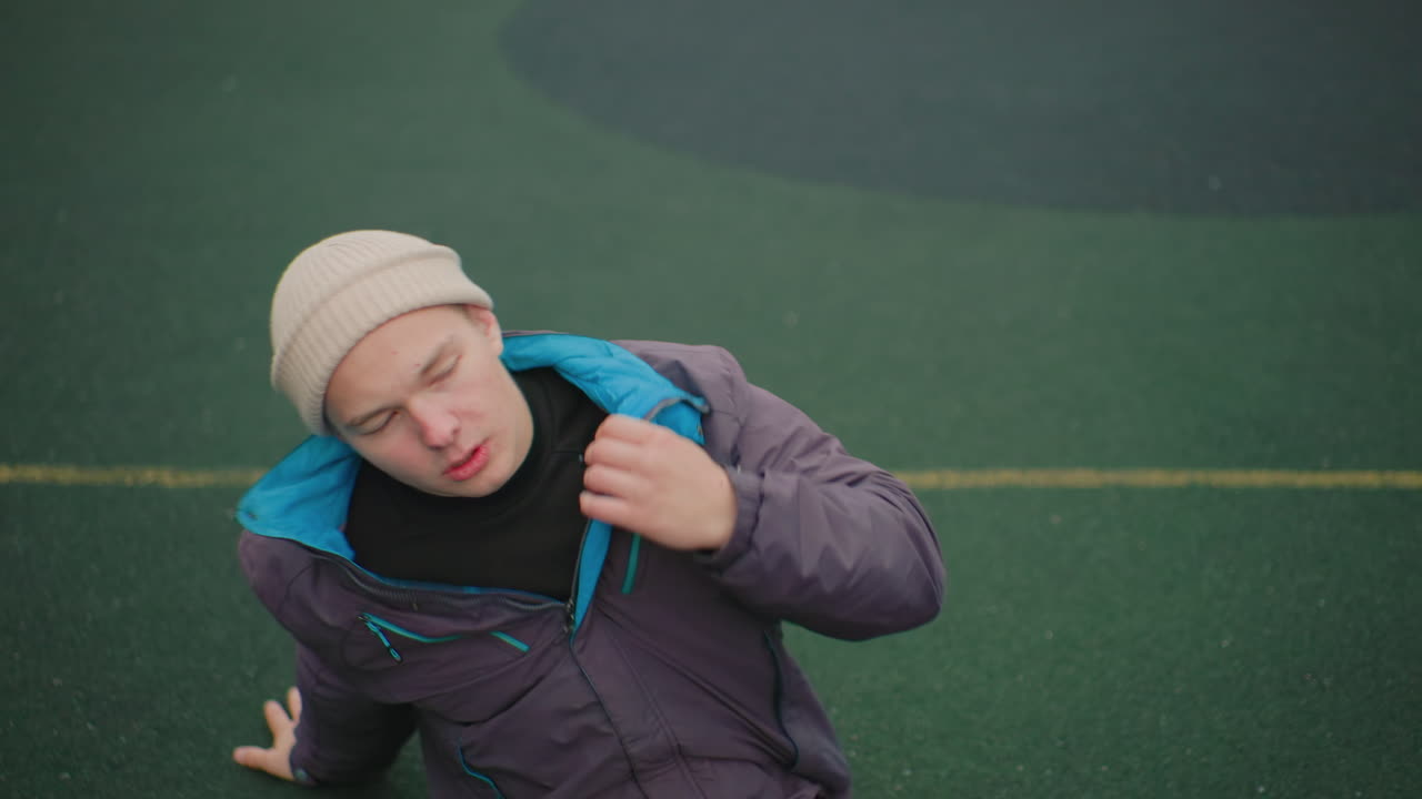 Sliten, hvit mann som sitter på banen med en basketball mellom beina, iført lue og vinterjakke. Han prøver å få tilbake pusten etter å ha spilt, kunstgress under føttene, overskyet himmel, urbant alene, ærlig portrett.