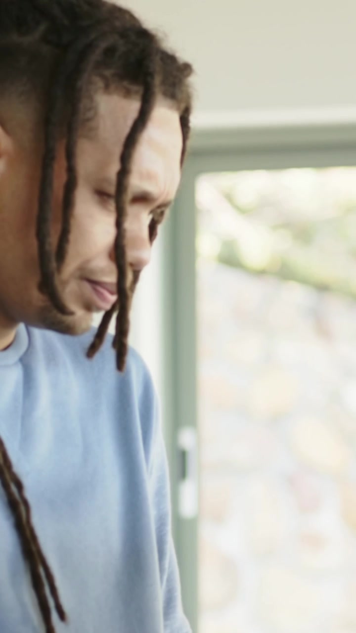Vídeo vertical: Homem com dreadlocks, de moletom, inclinado em casa, segurando e consertando um laptop.