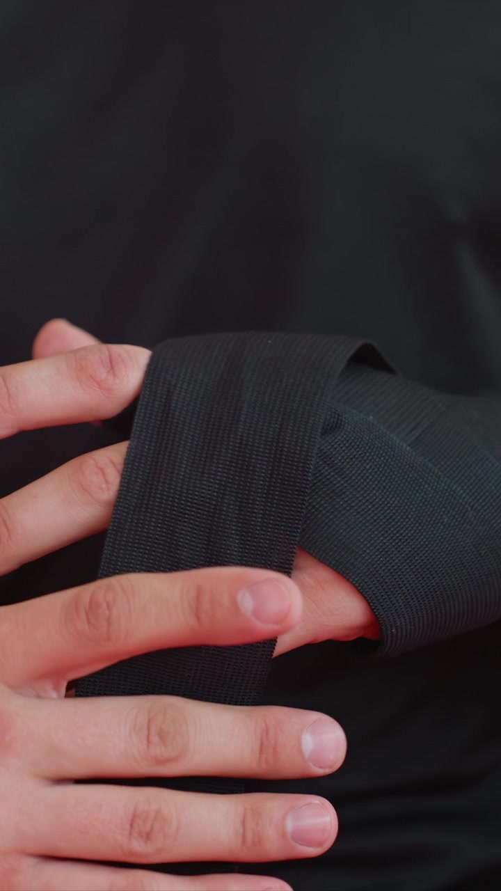 Close up of hands wrapping black material around fingers and wrist. Hands manipulate fabric securing it tightly. Dark background emphasizes hand actions in controlled environment