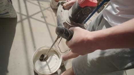 mixing of construction materials, with a worker using a power drill and mixing paddle to blend plaster or paint in a bucket, a common practice in building and renovation work