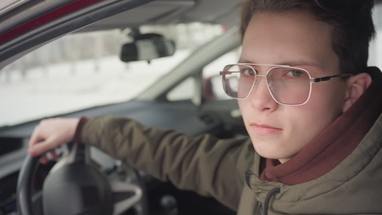 close up of boy seated in driver seat rolling down window while holding steering wheel with soft elegant expression, blurred snowy background and raindrops on glass creating wintery atmosphere