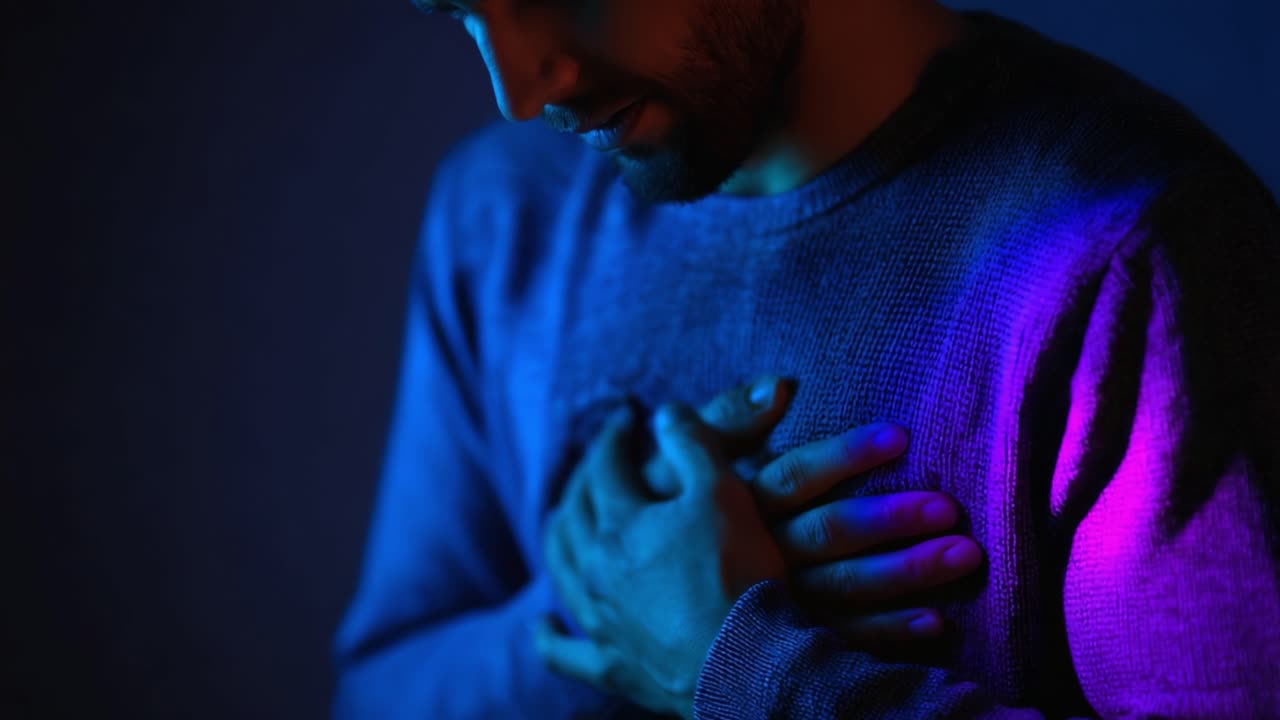 A man in a dark atmosphere places his hand over his chest, highlighting an emotional moment illuminated by soft purple and blue lights, conveying deep introspection and sensitivity