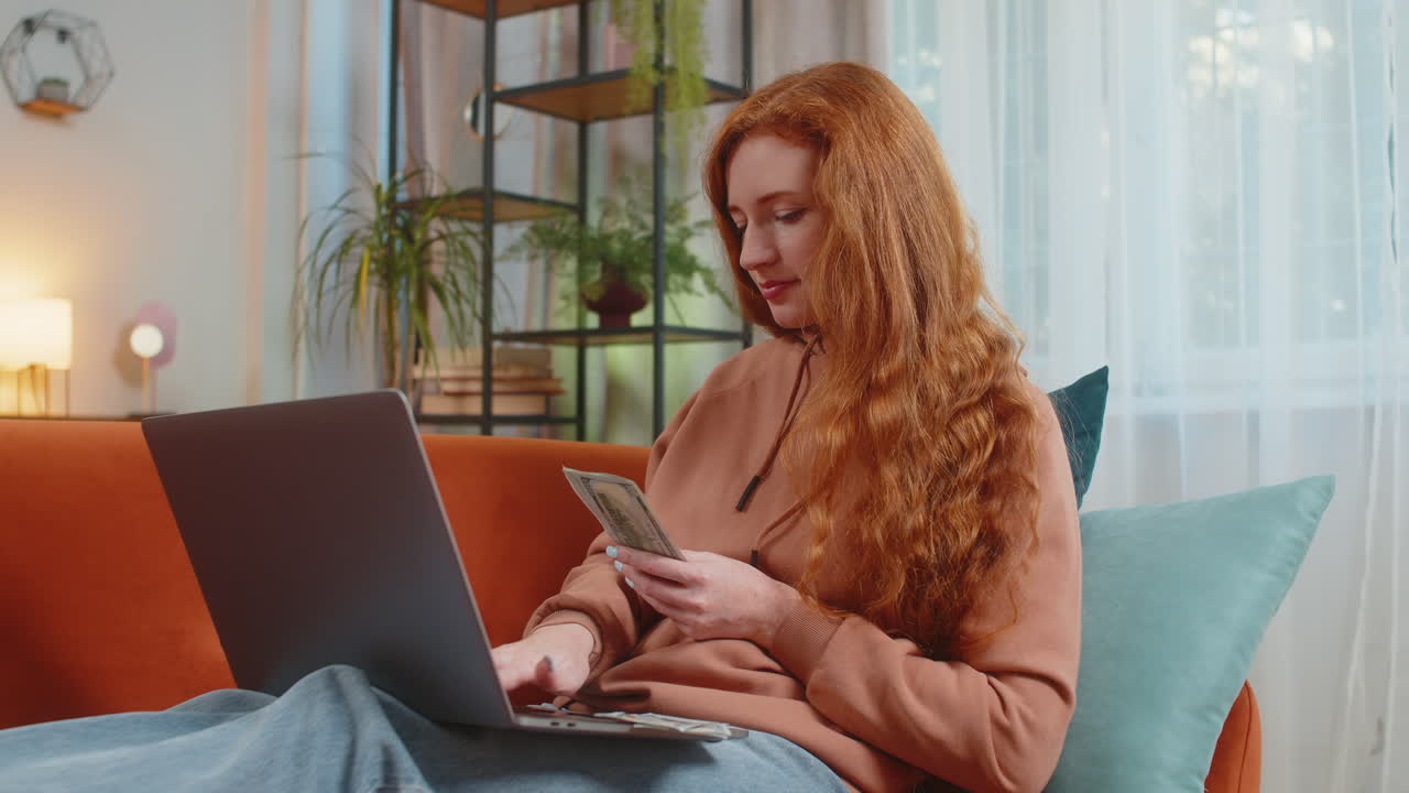 Rich happy redhead woman counting money cash using laptop calculate domestic bills sitting on couch