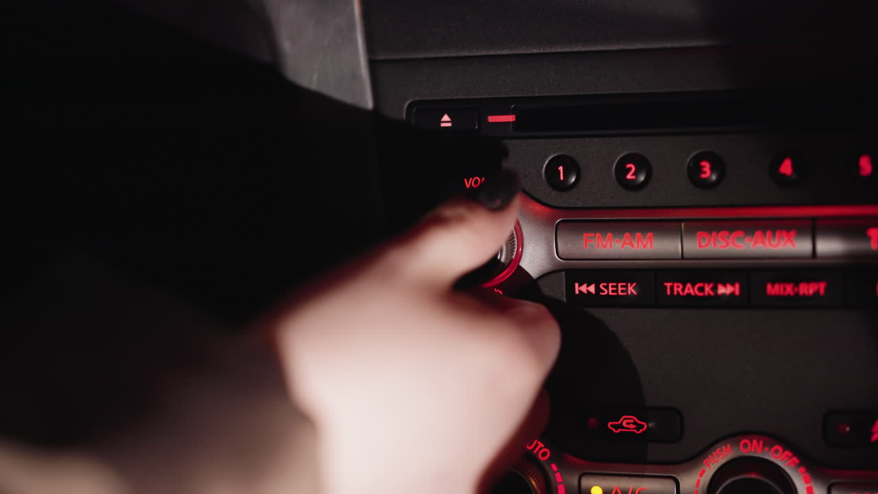 vista en primera persona de la mano de una mujer girando el botón de volumen de la radio fm dentro de un coche por la noche. el tablero de instrumentos está iluminado con luces rojas, destacando los controles de radio. disparado con una cámara de mano