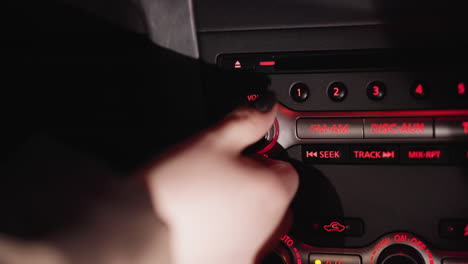 first-person view of a woman's hand turning the volume knob on the fm radio inside a car at night. the dashboard is illuminated with red lights, highlighting the radio controls. shot with a handheld camera