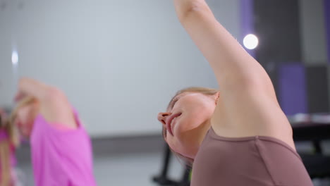 primer plano de mujeres sentadas en esteras realizando ejercicios de estiramiento de costillas en un gimnasio bien equipado, el fondo presenta equipos de entrenamiento borrosos, espejos y un entorno de fitness moderno