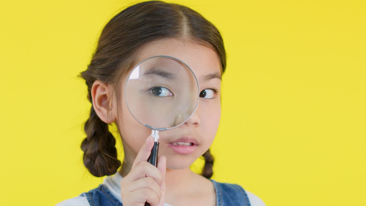 Young girl with braids distorts her face using magnifying glass against bright yellow background, close-up
