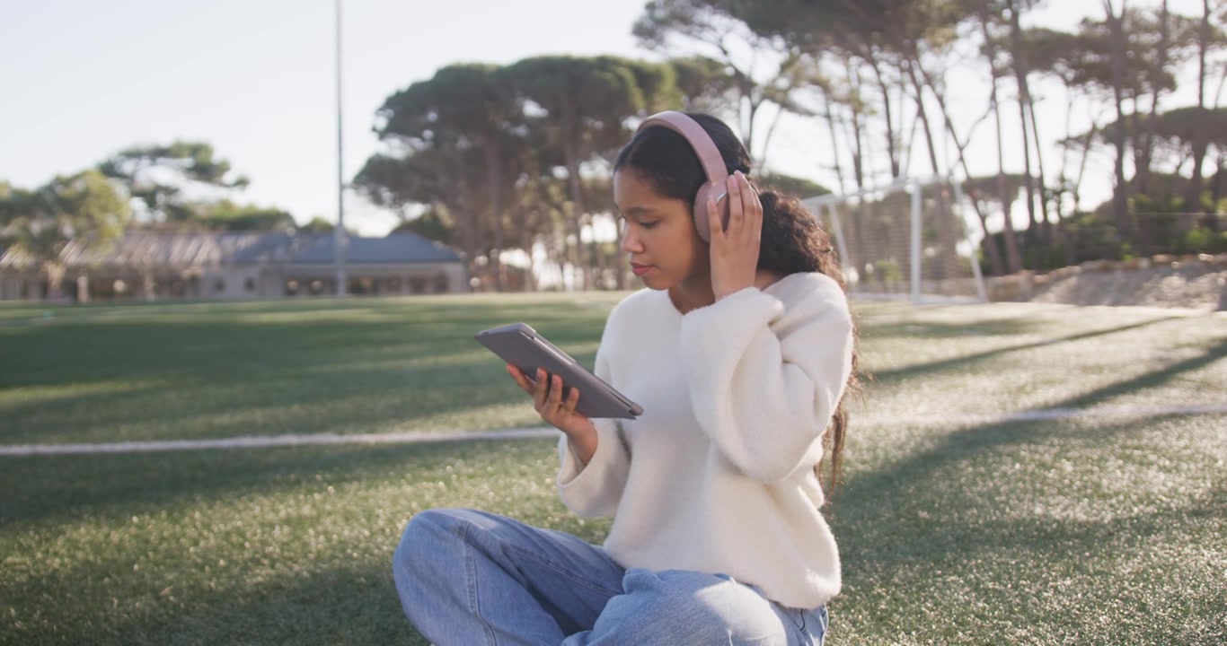 Ragazza adolescente seduta sul prato con il tablet, che sistema le cuffie rosa e ascolta dopo aver toccato "play", con spazio per il testo.