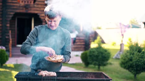 young man stands on the background of a summer house lays meat on barbecue grill