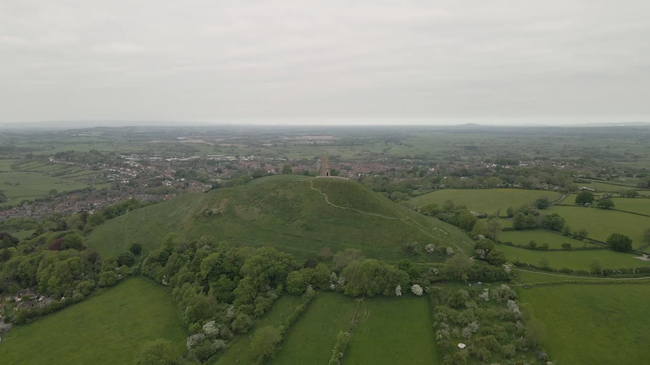 Aerial view of Glastonbury, drone rotating to te left side shoeing the tower and the extension of Glastonbury, drone flying over the green fields . 4K, 60fps.