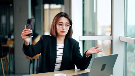 caucasian woman wearing glasses holding her phone on the face level. business lady talks via video chat pointing at her laptop with a hand.