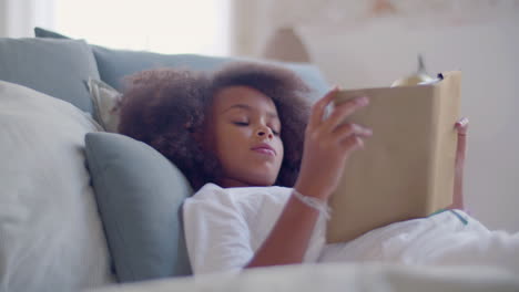 dolly medium shot of a litlle african american girl in white pyjamas lying in bed and reading book