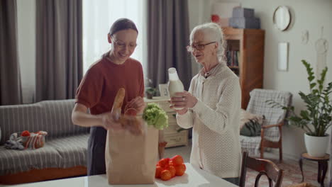 young woman helping elderly grandmother unpack groceries at home
