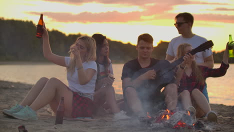 los estudiantes tocan la guitarra y cantan canciones alrededor de la fogata en la fiesta de la playa. esta es una conmovedora noche de verano con los mejores amigos y la cerveza.