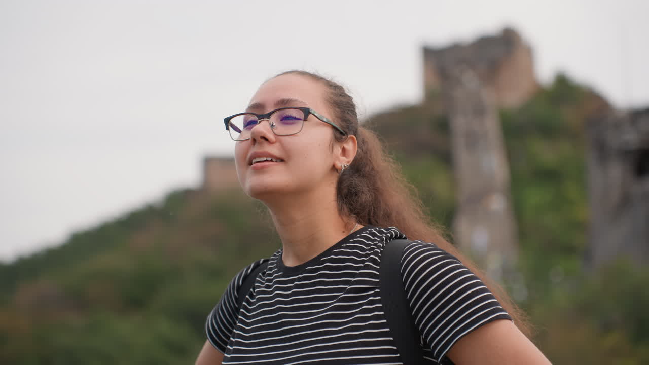 Thoughtful young woman admiring ruins in closeup portrait, glasses catching soft light contemplative student or history enthusiast looking toward stone towers, serene hillside background, heritage
