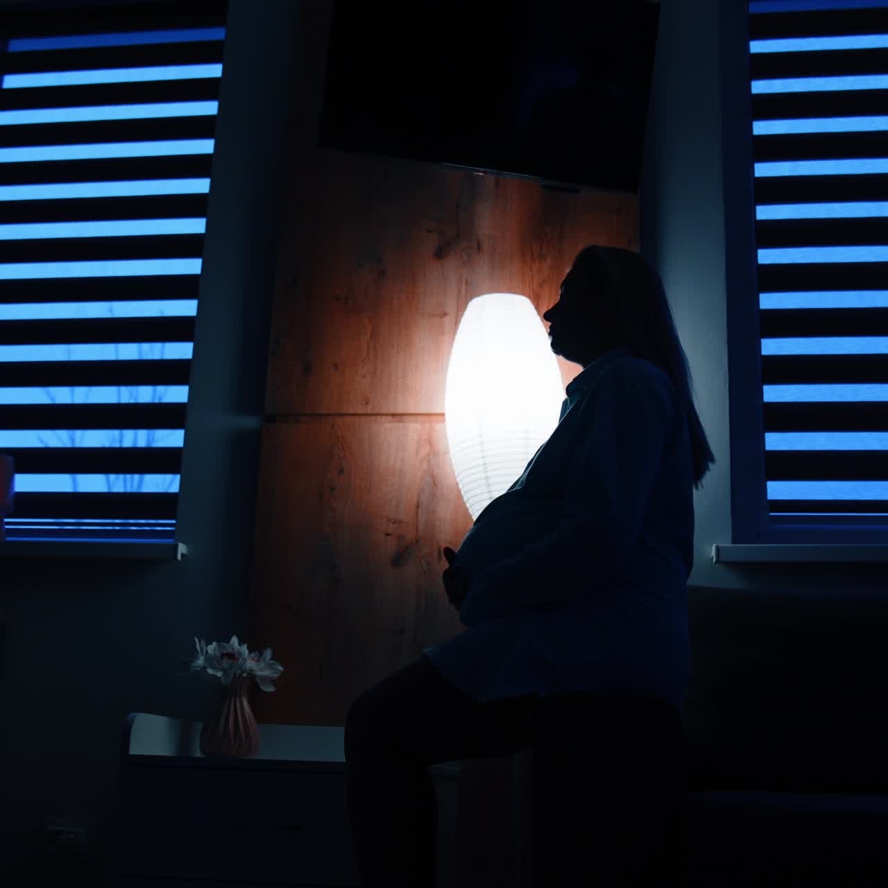 Calm long-haired pregnant woman sits on a sofa handrail in dark room. Future mom strokes her belly with love. Low angle view