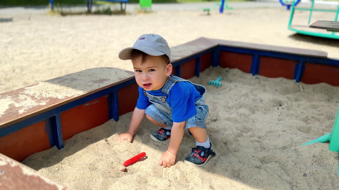 Tiny baby boy plays in a sandbox. Child holds his hands in the sand but looks around.